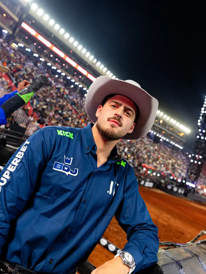 Jon na arena da PBR, de chapéu, com a arquibancada ao fundo