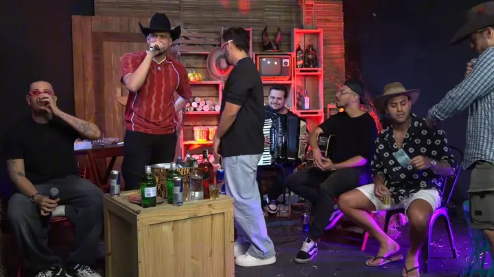 Convidado canta ao microfone no cenário do bar, com a roda de amigos sentada em volta